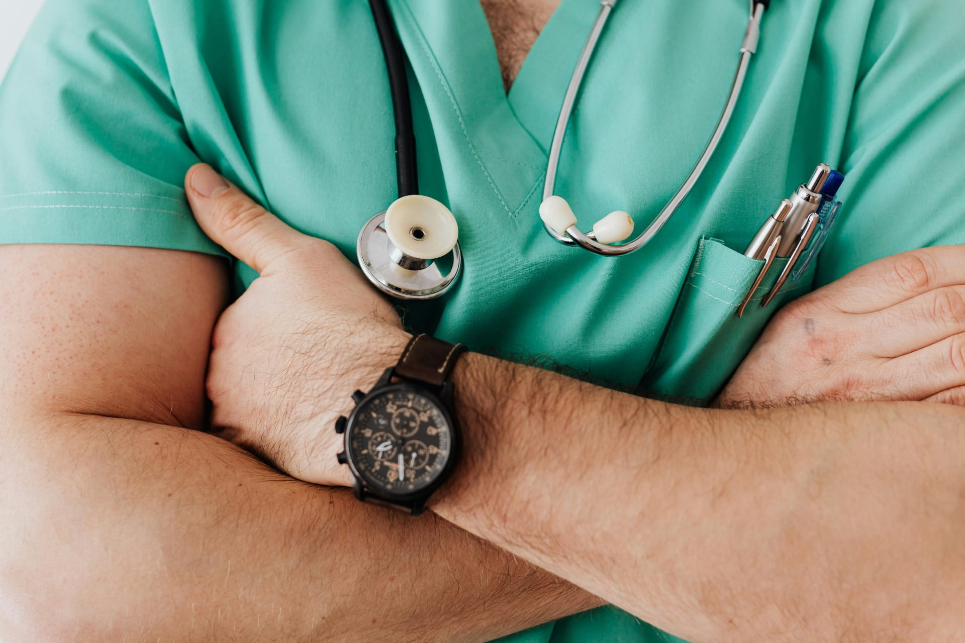 Manos con estetoscopio y bata verde quirúrgica, reloj de pulsera y herramientas médicas visibles. Seguro Salud Empresas