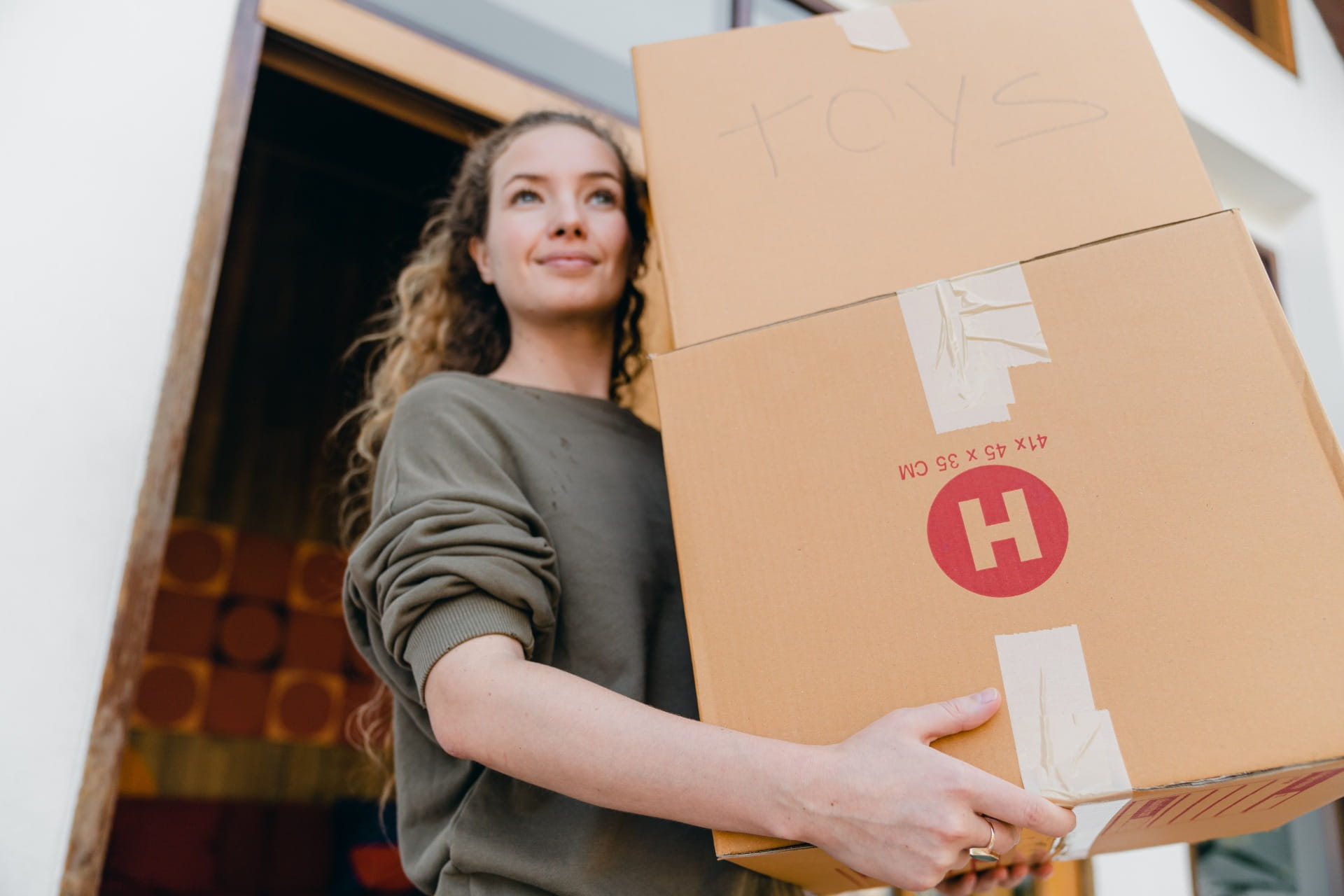 Mujer sonriente sostiene una caja de cartón grande con el logo rojo H, en la entrada. Seguro de transporte de carga, terrestre, marítimo, aéreo