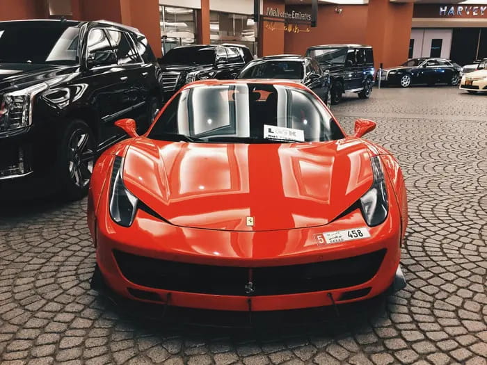 Coche deportivo rojo con capó y faros delanteros, estacionado en un estacionamiento con otros autos alrededor. Seguro para vehículos, autos, motos