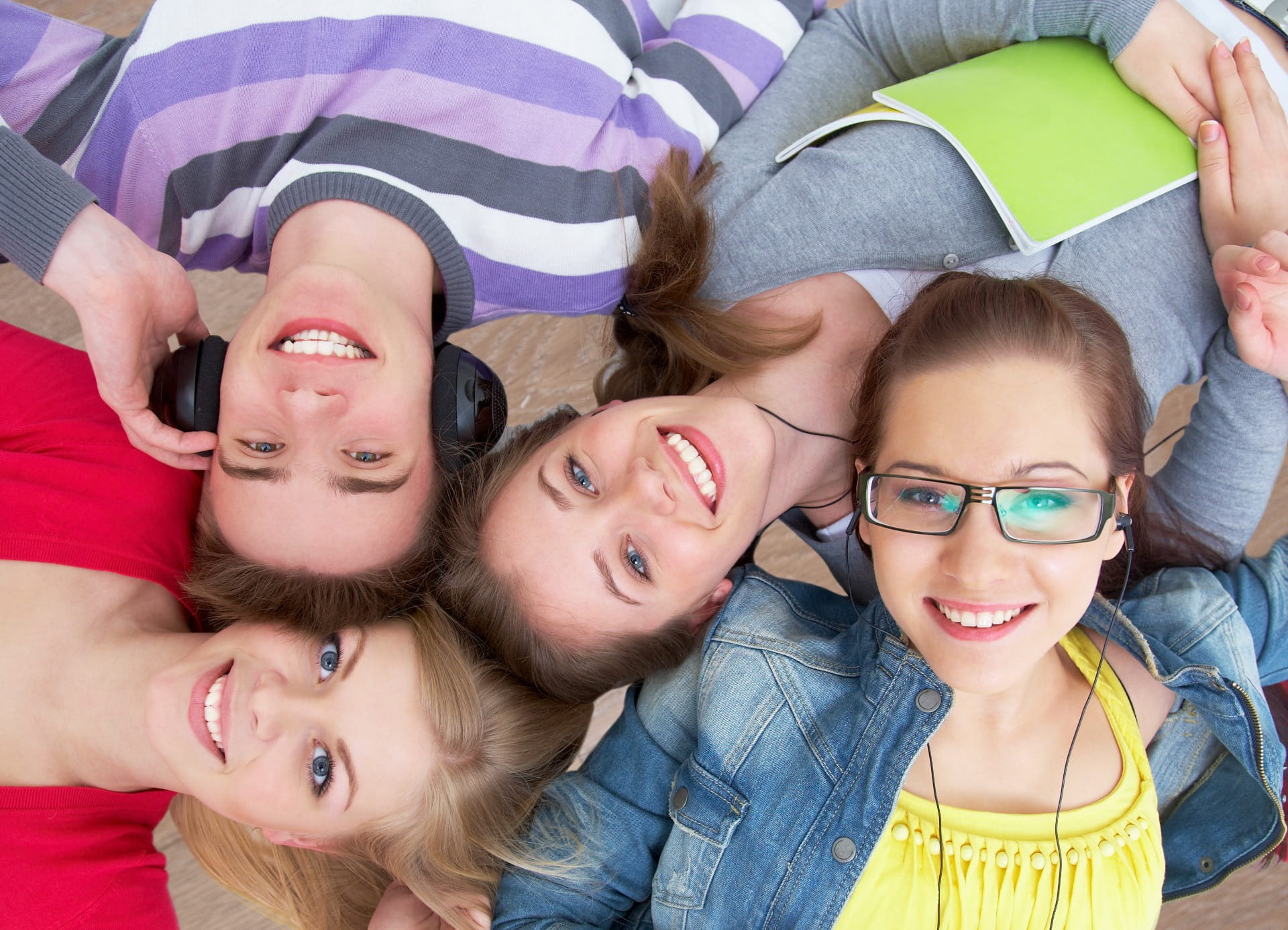 Cuatro jóvenes sonrientes tumbados en el sofá, con auriculares y un cuaderno verde abierto. Seguro de salud complementario individual.