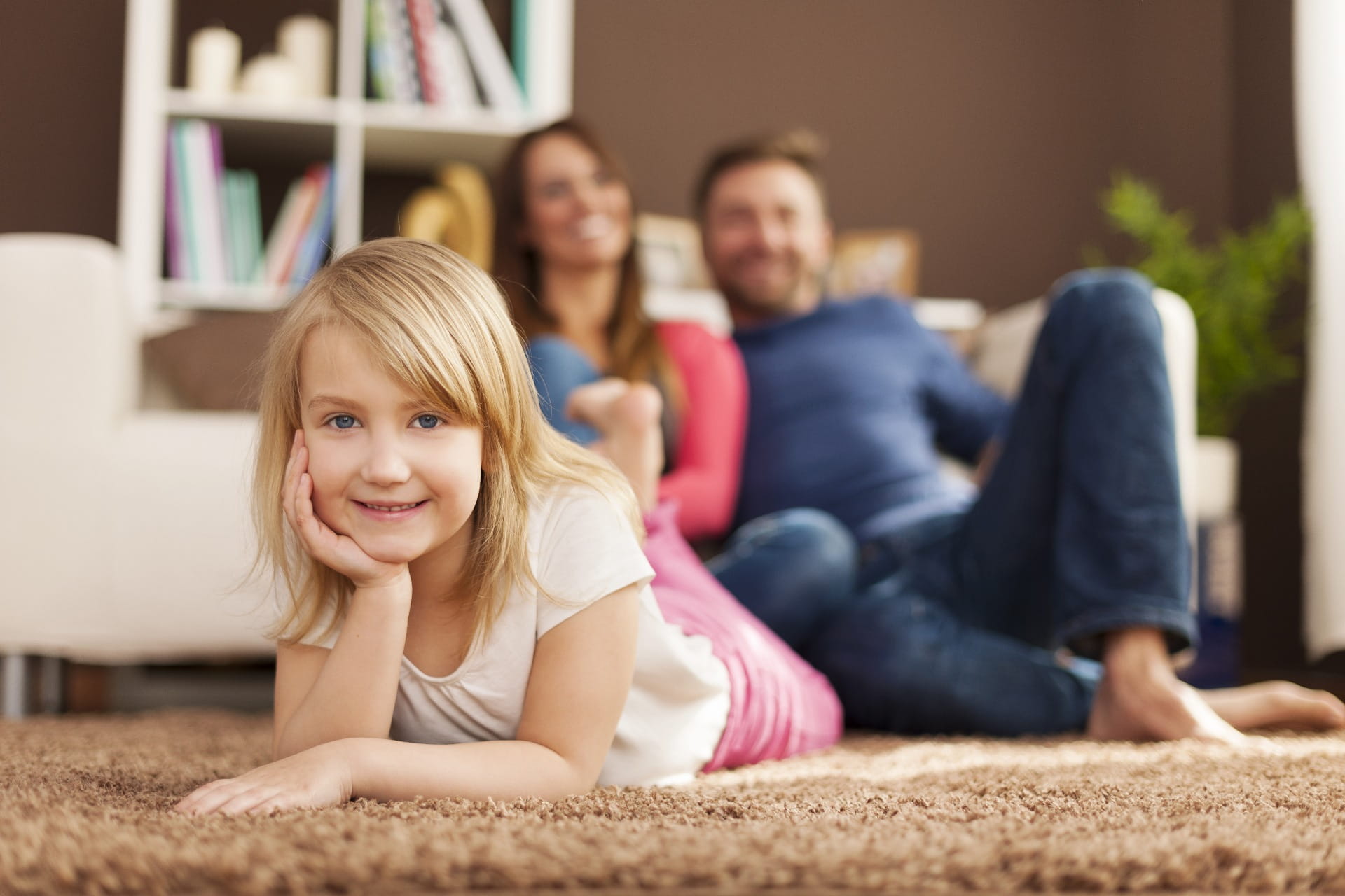 Niña rubia sonriente en primer plano, con familia sentada al fondo en una sala acogedora. Seguros de Vida